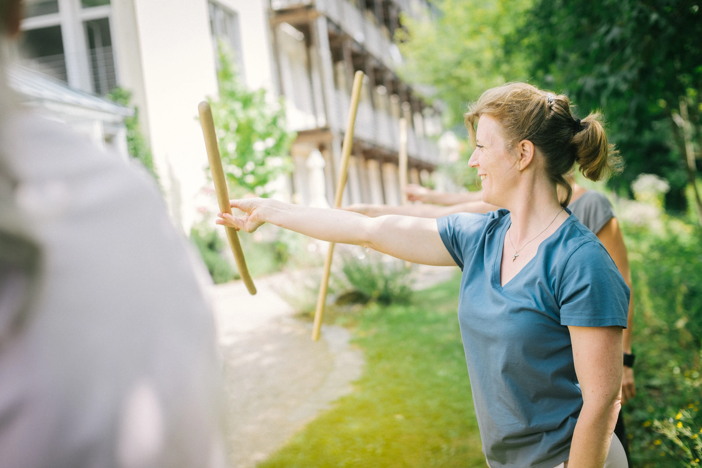Frau macht lächelend übungen draußen in einer gruppe mit holz stöcken